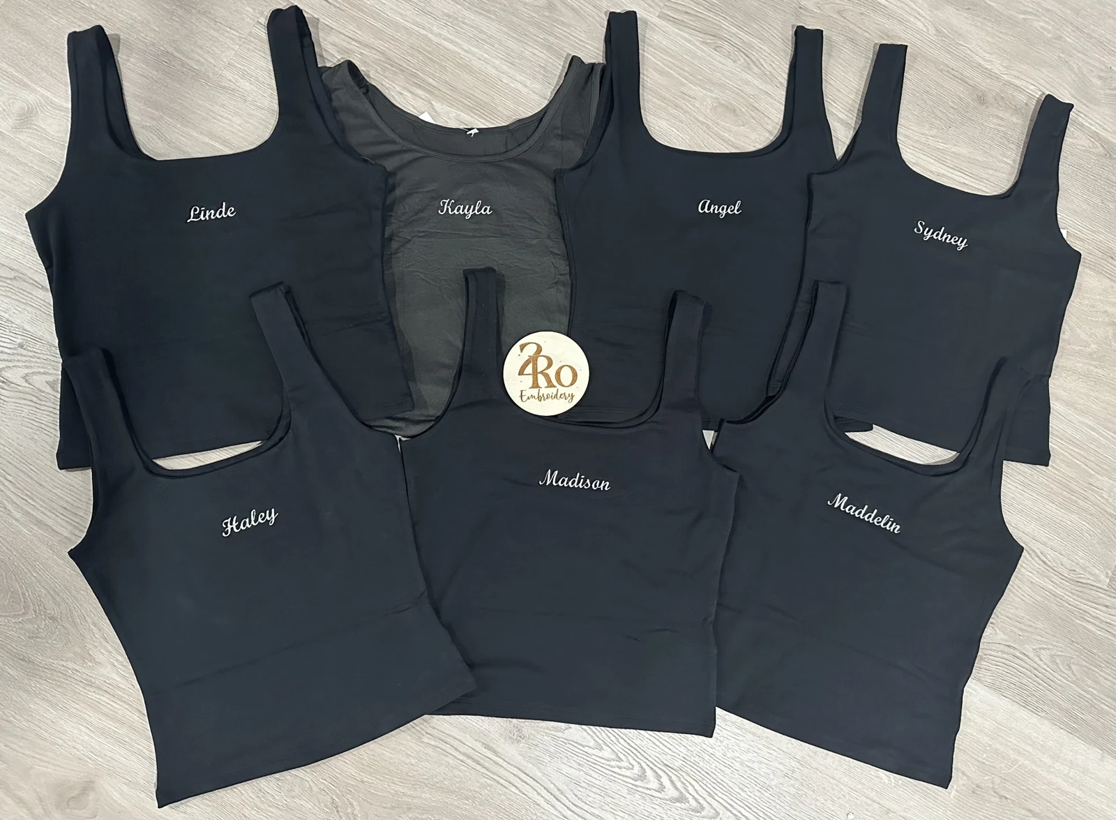 Seven black tank tops, each embroidered with a different name in white script