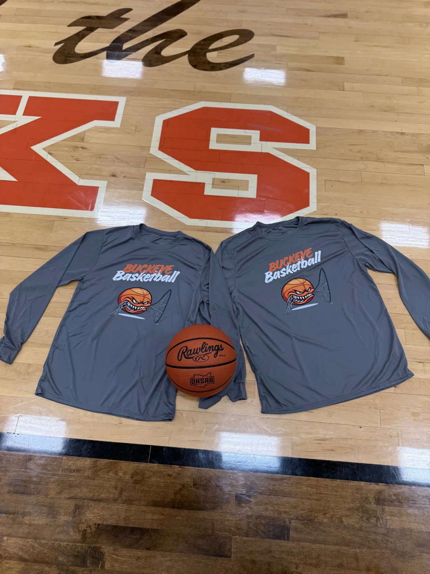 Two gray long-sleeve shirts printed with Buckeye Basketball and a fierce cartoon basketball, on a gym floor