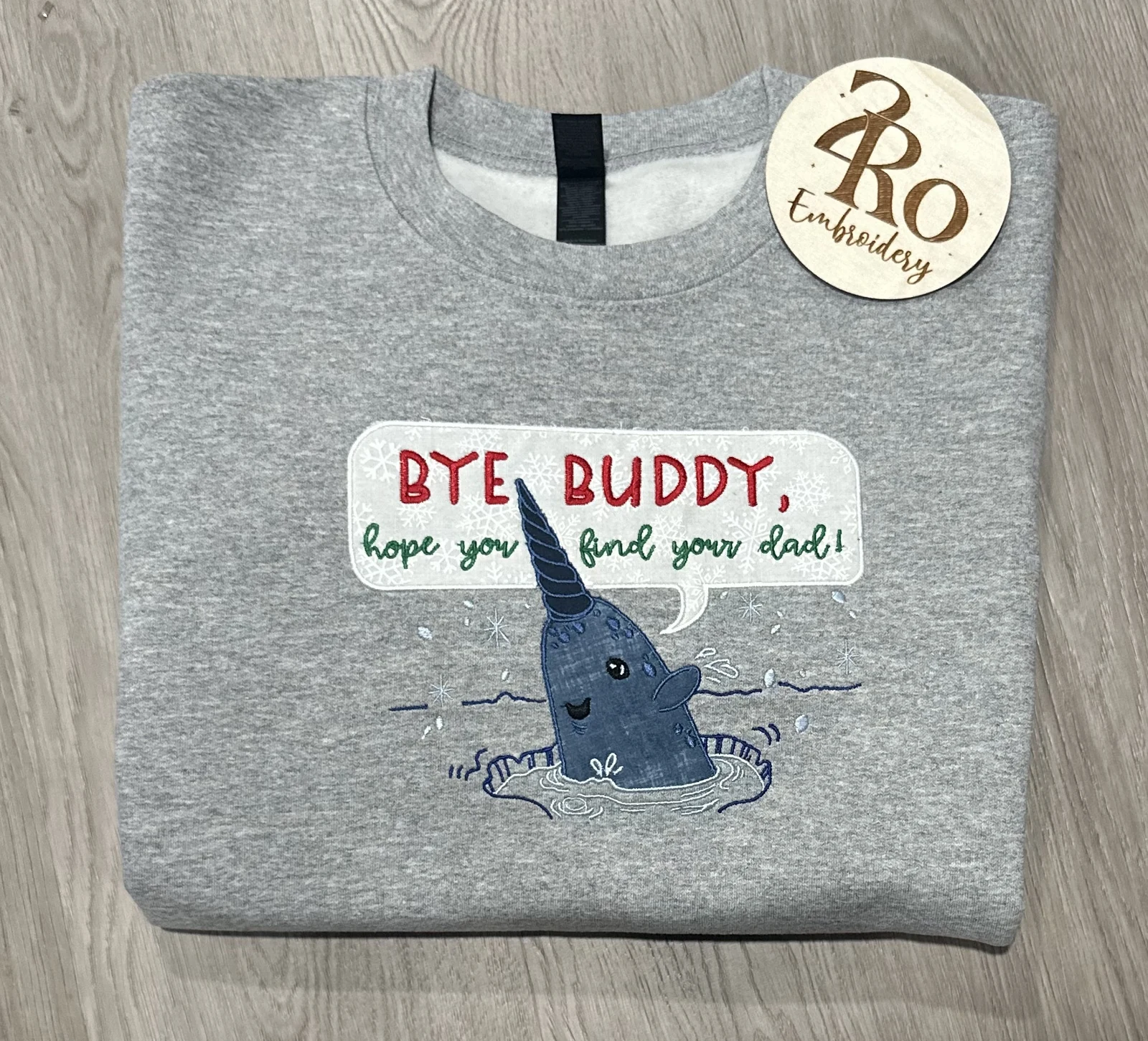 Gray sweatshirt with an appliqué narwhal and a snowflake speech bubble reading Bye buddy, hope you find your dad