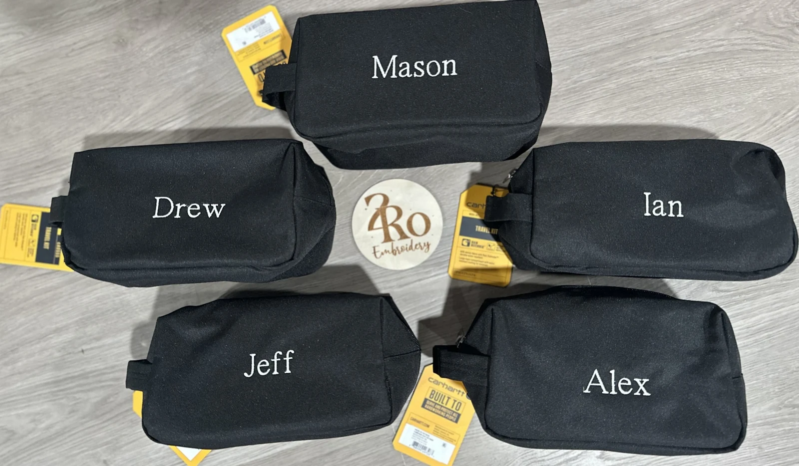 Five black travel kits, each embroidered with a different name in white