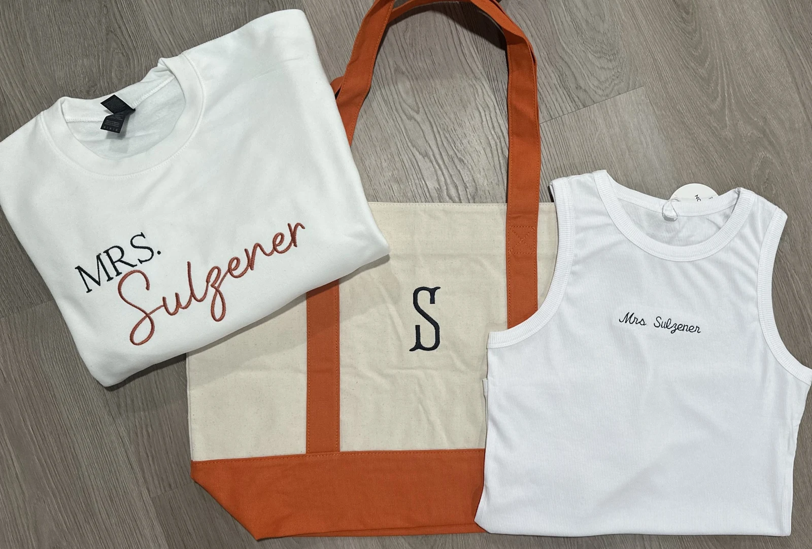 White sweatshirt, canvas tote with an orange trim and initial, and white tank top, each embroidered with a married name