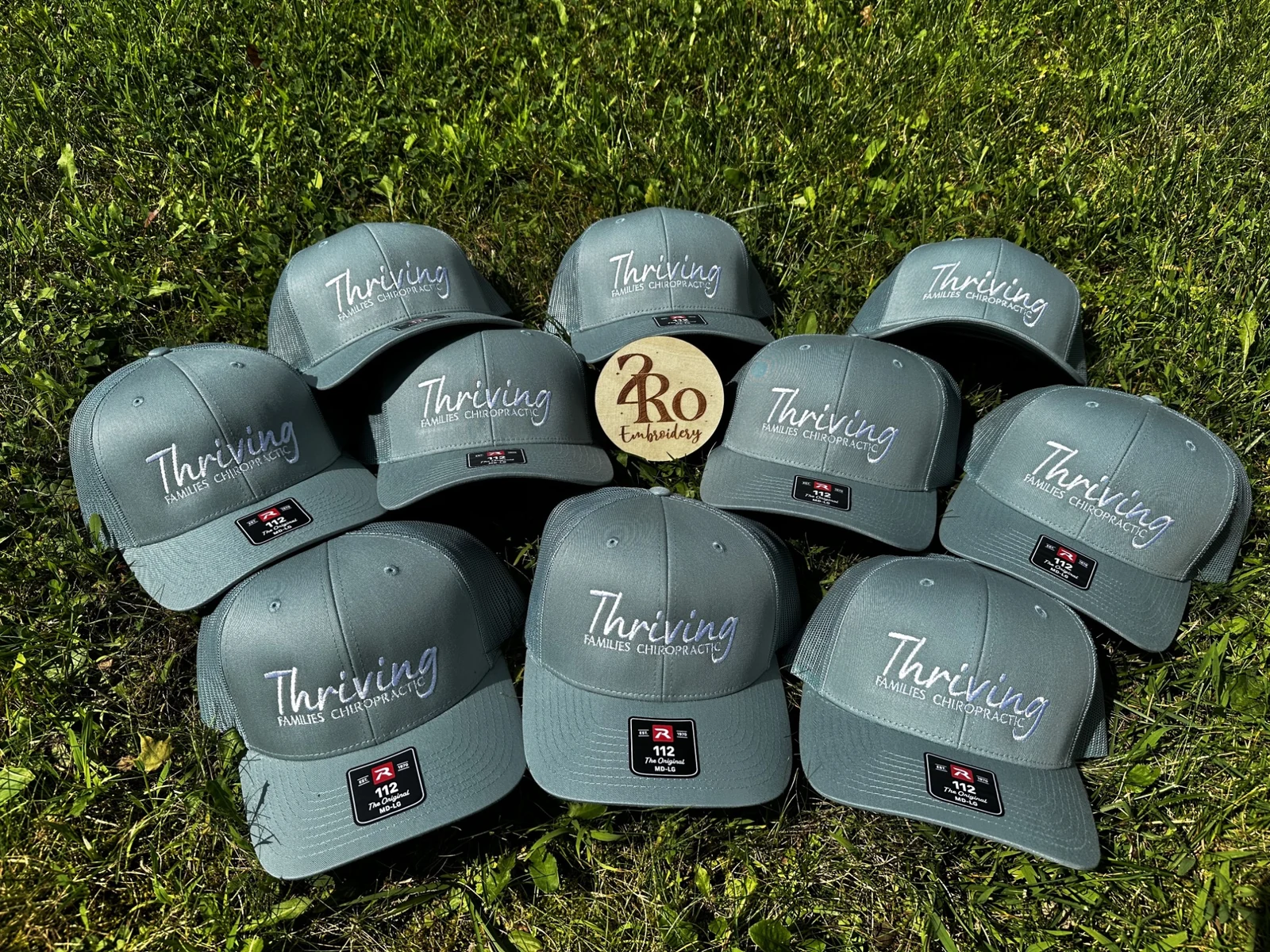 Sage trucker hats embroidered with the white Thriving Families Chiropractic logo, laid out on grass