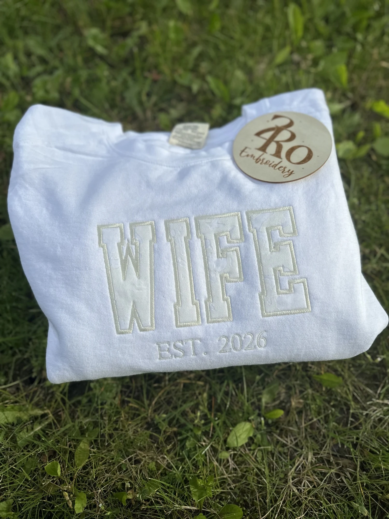 White sweatshirt with cream appliqué WIFE lettering and EST. 2026, on grass